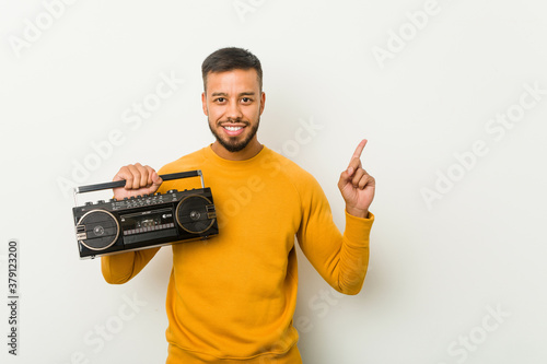 Young south-asian man holding a guetto blaster smiling cheerfully pointing with forefinger away.