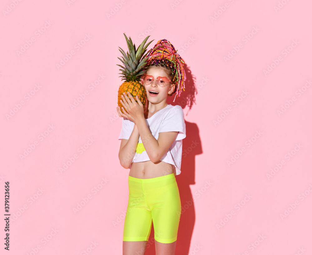 Active girl with rainbow colored african braids on head holding pineapple and pointing at camera with cheerful smile. Cropped shot isolated on pink