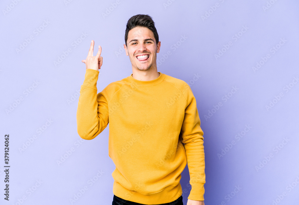 Young caucasian man isolated on purple background showing rock gesture with fingers Young caucasian man isolated on purple background showing rock gesture with fingers