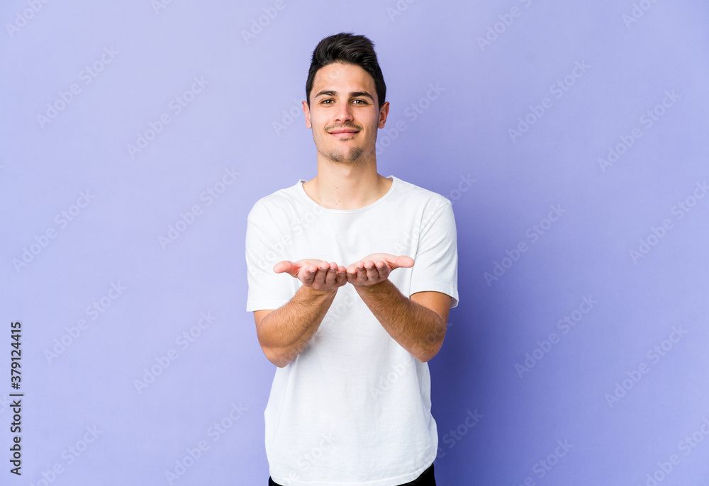 Young caucasian man isolated on purple background holding something with palms, offering to camera.