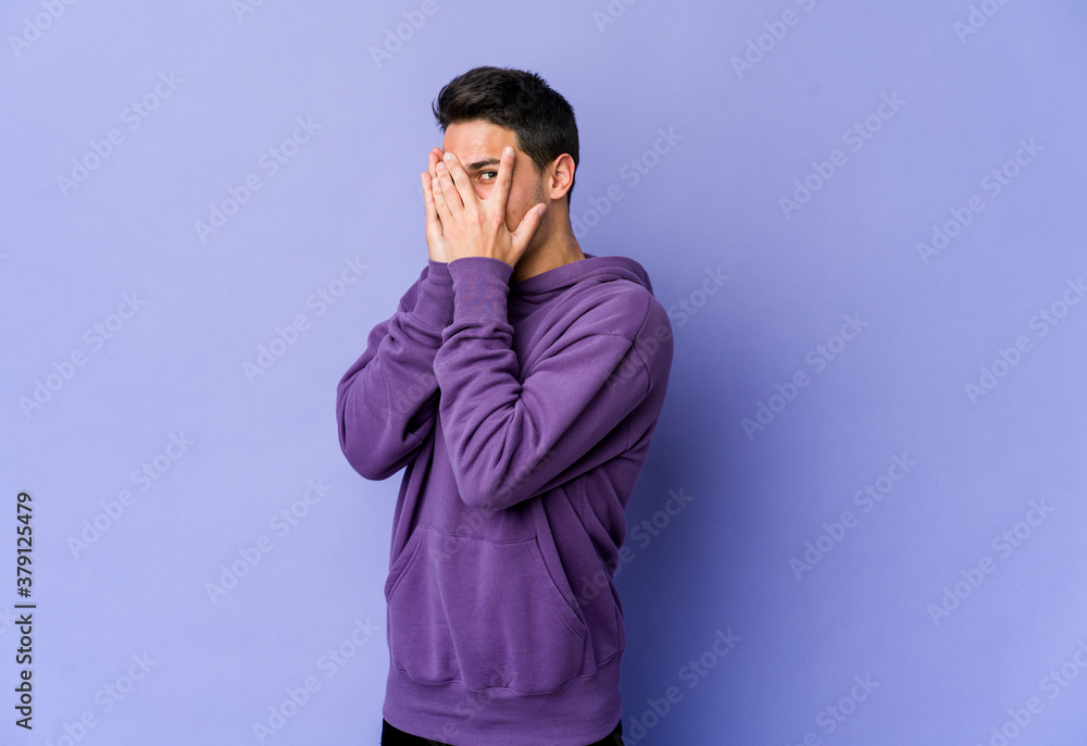 Young caucasian man isolated on purple background blink through fingers frightened and nervous.