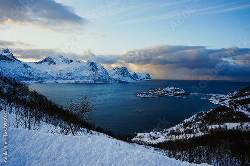 A fishing village picturesquely situated on the island. Beautiful winter scenery. Amazing view from the hill. Natural norwegian landscape. Senja island, north Norway. Snowy mountains and arctic sea.