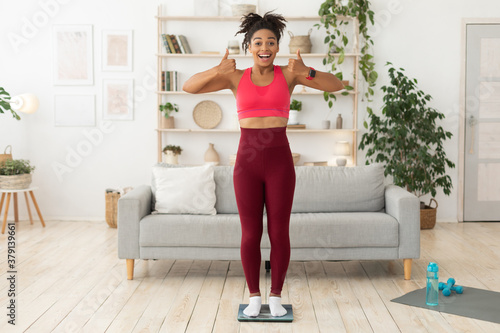 Black Fitness Lady Gesturing Thumbs-Up Standing On Scales At Home