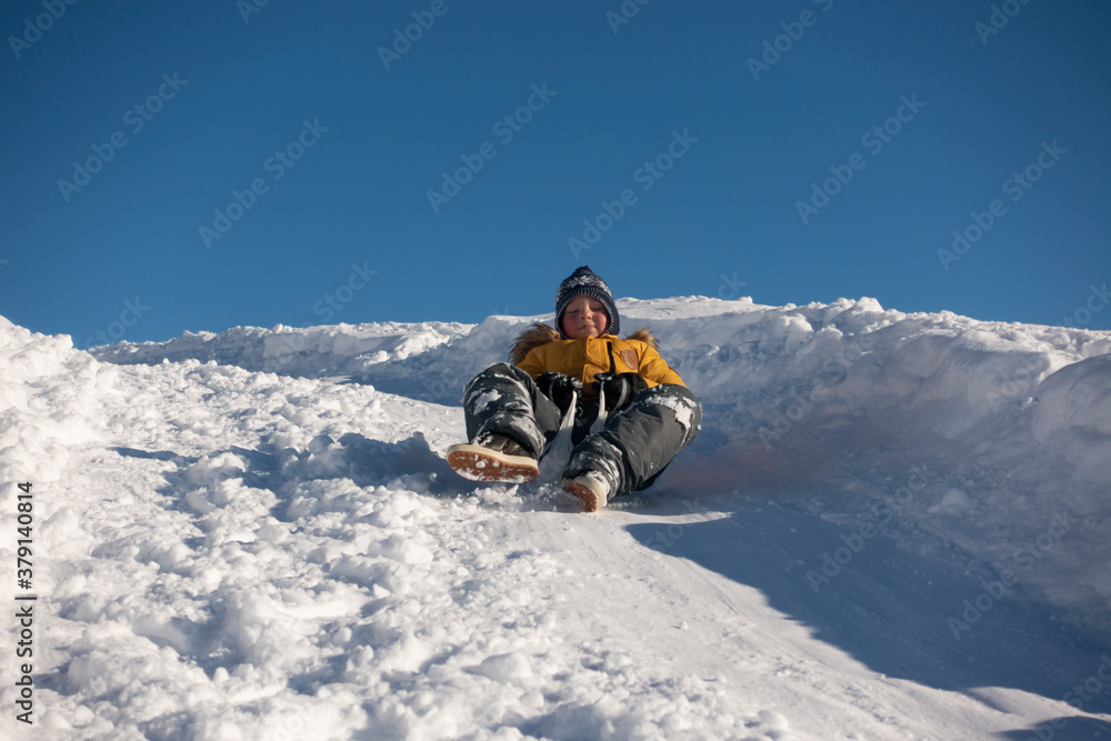 happy boy sliding down snow hill on sled outdoors in winter, cold season concept