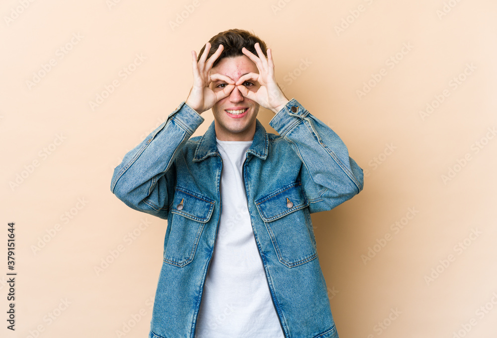 Young caucasian man isolated on beige background showing okay sign over eyes Young caucasian man isolated on beige background showing okay sign over eyes