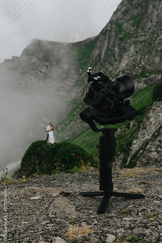 stabilizer with a camera standing in the mountains in cloudy weather, behind him is the groom and the bride in a wedding dress, wedding photography