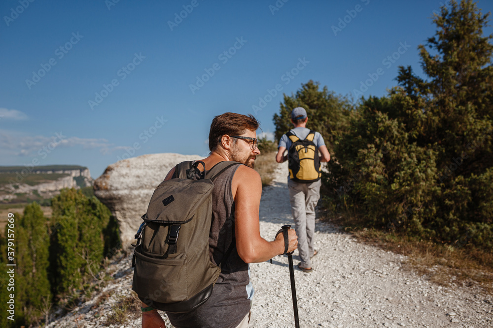 Two Men walking on rocky slope carrying Backpacks using trekking Sticks.