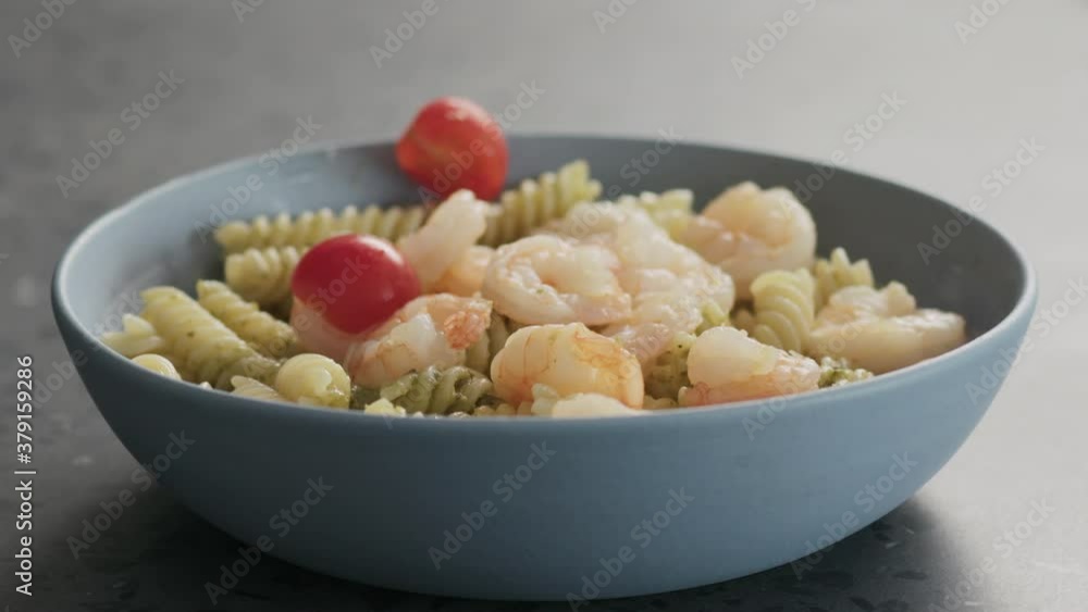 Slow motion add tomatoes to pesto fusilli pasta with shrimps in blue bowl on concrete background Slow motion add tomatoes to pesto fusilli pasta with shrimps in blue bowl on concrete background