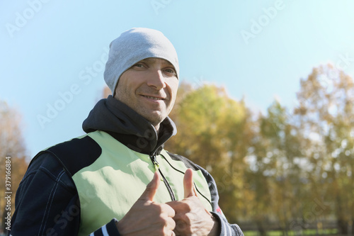 middle aged sportsman showing thumbs up and smiling. man getting rest after jogging. handsome man wearing sport jacket and a beanie hat on autumn background. cold sason outdoors activity. autumn sport photo