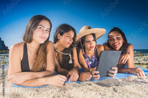 Four latin american ethnic girls friends using social media in digital tablet lying on sand Caribbean beach Los Roques
