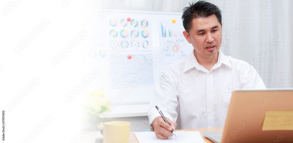 Obraz premium Asian businessman talking with team via video conference writing note on paper. business people using laptop for online meeting. Business work from home concept.