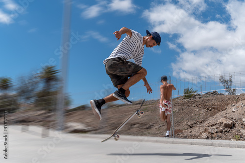 Hombre joven hace un salto con su tabla en un parque de skateboard en un dia soledado. En el fondo, otro hombre hace estiramientos photo