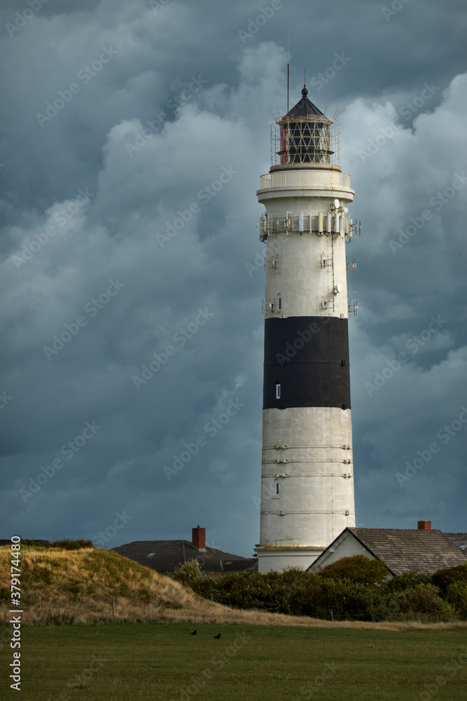 White lighthouse with a black stripe behind a meadow and houses in front of a dramatic grey cloudy sky on Sylt, Germany