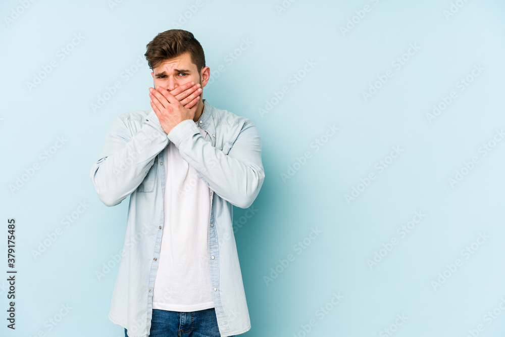 Young caucasian man isolated on blue background covering mouth with hands looking worried.