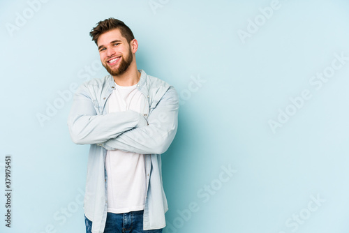 Young caucasian man isolated on blue background who feels confident, crossing arms with determination. © Asier