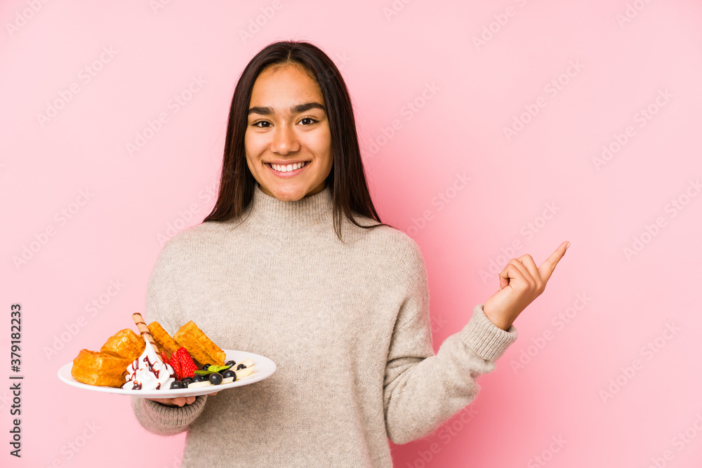 Young asian woman holding a waffle isolated smiling and pointing aside, showing something at blank space. Young asian woman holding a waffle isolated smiling and pointing aside, showing something at blank space.