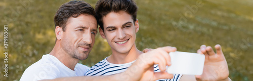 panoramic crop of father and teenager son taking selfie in park