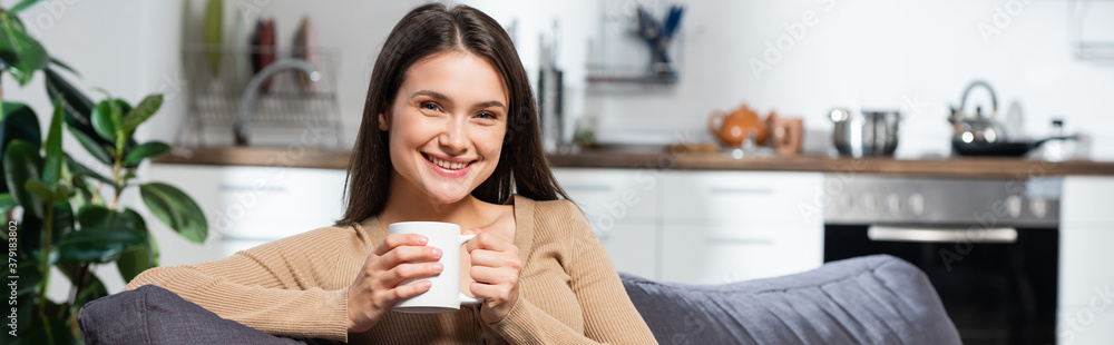 horizontal concept of pleased woman looking at camera while sitting on couch in kitchen with cup of warming beverage horizontal concept of pleased woman looking at camera while sitting on couch in kitchen with cup of warming beverage