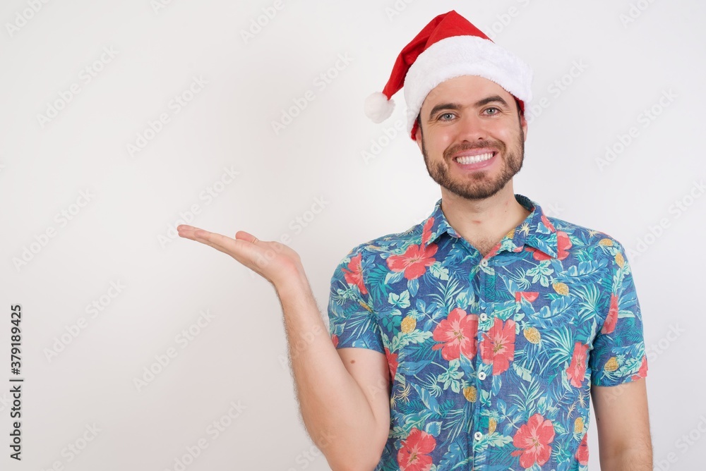 Young caucasian man wearing hawaiian shirt and Santa hat over isolated white background smiling cheerful presenting and pointing with palm of hand looking at the camera. Young caucasian man wearing hawaiian shirt and Santa hat over isolated white background smiling cheerful presenting and pointing with palm of hand looking at the camera.