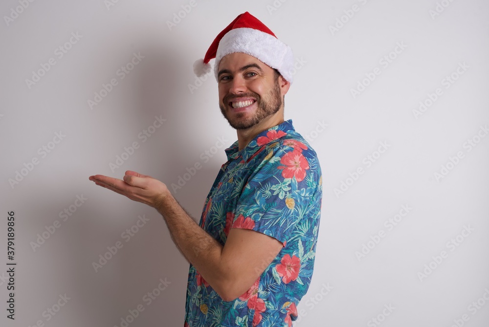 Young caucasian man wearing hawaiian shirt and Santa hat over isolated white background pointing aside with hands open palms showing copy space, presenting advertisement smiling excited happy