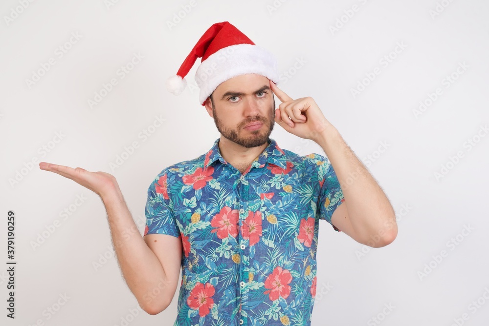 Young caucasian man wearing hawaiian shirt and Santa hat over isolated white background confused and annoyed with open palm showing copy space and pointing finger to forehead. Think about it. Young caucasian man wearing hawaiian shirt and Santa hat over isolated white background confused and annoyed with open palm showing copy space and pointing finger to forehead. Think about it.
