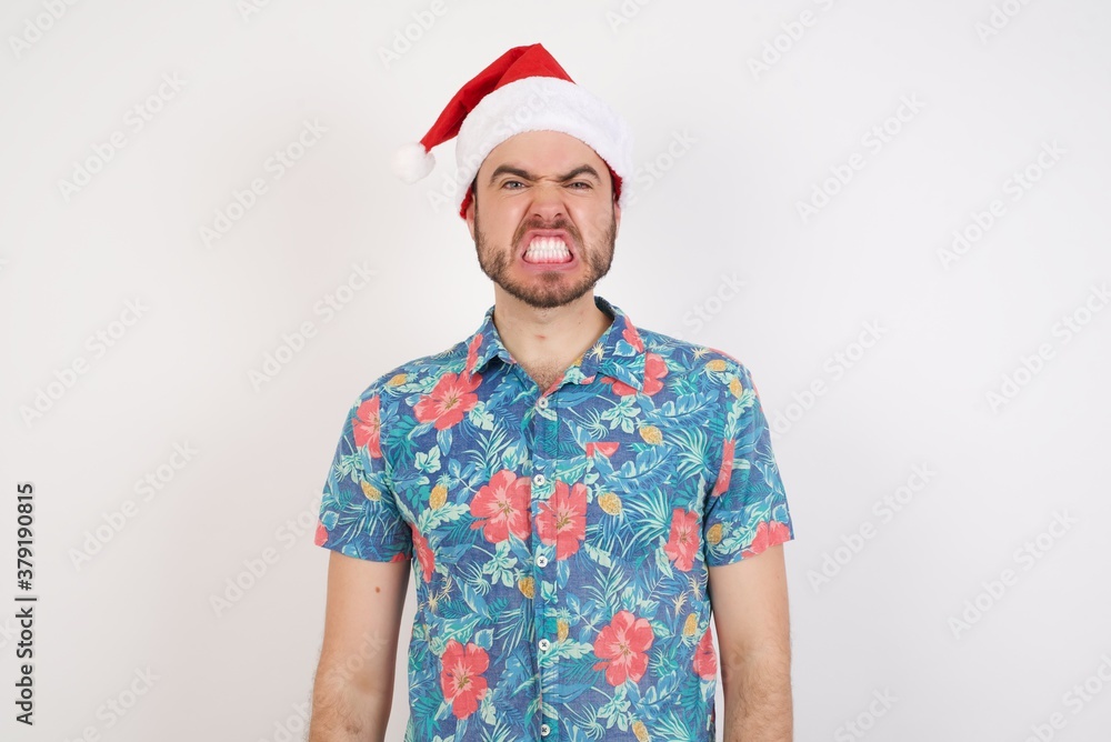 Angry Young caucasian man wearing hawaiian shirt and Santa hat over isolated white background, keeps teeth clenched, frowns face in dissatisfaction, irritated because of much duties. Furious man Angry Young caucasian man wearing hawaiian shirt and Santa hat over isolated white background, keeps teeth clenched, frowns face in dissatisfaction, irritated because of much duties. Furious man