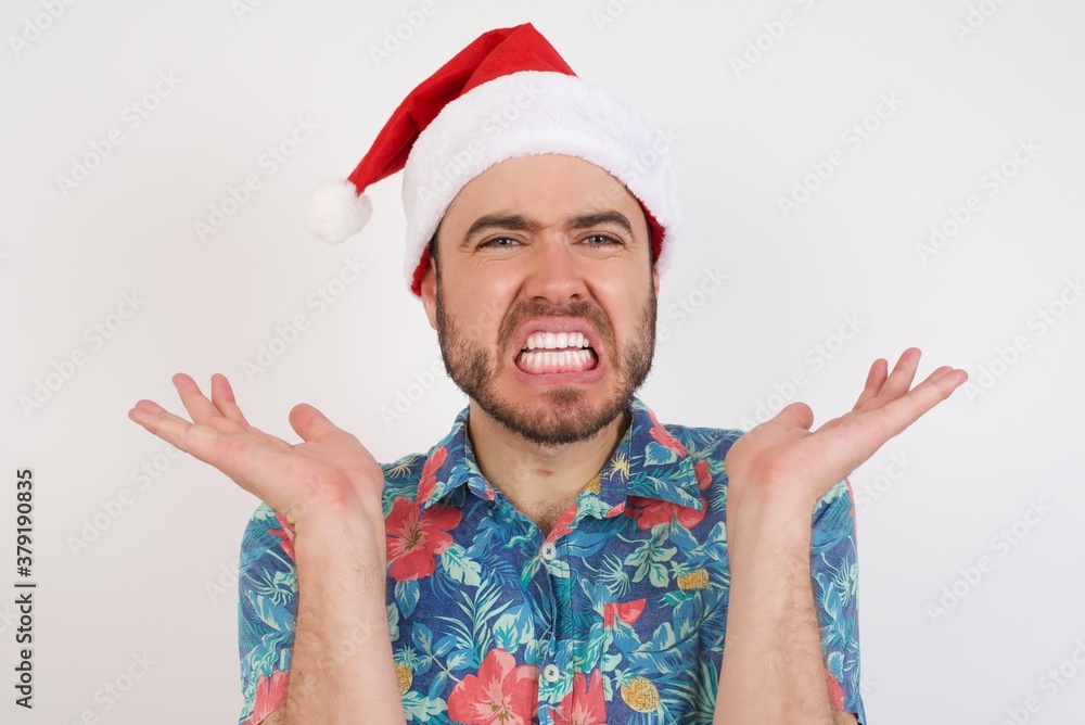 Indignant Young caucasian man wearing hawaiian shirt and Santa hat gestures in bewilderment, frowns face with dissatisfaction, says that she forgot to buy products for preparing supper. Indignant Young caucasian man wearing hawaiian shirt and Santa hat gestures in bewilderment, frowns face with dissatisfaction, says that she forgot to buy products for preparing supper.