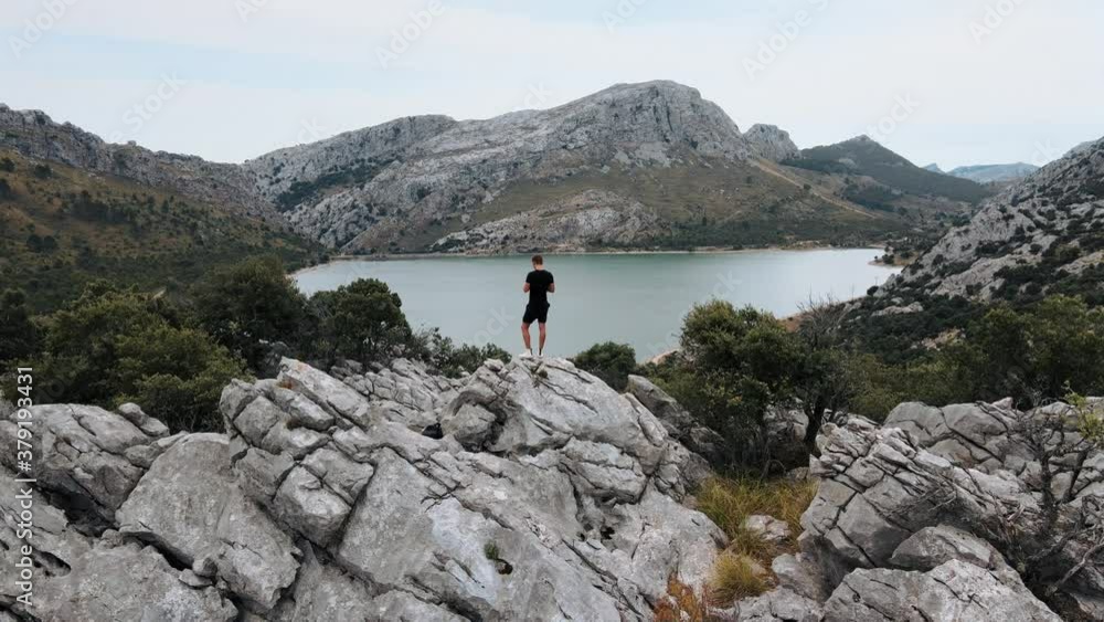 Aerial orbit shot of a man standing on a rocky hill overlooking a beautiful lake Aerial orbit shot of a man standing on a rocky hill overlooking a beautiful lake