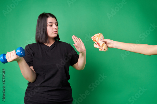 Fat young woman refuses junk food. Hand is offering unhealthy food. American thick lady with blue dumbbell is making no gesture. Isolated on green background.