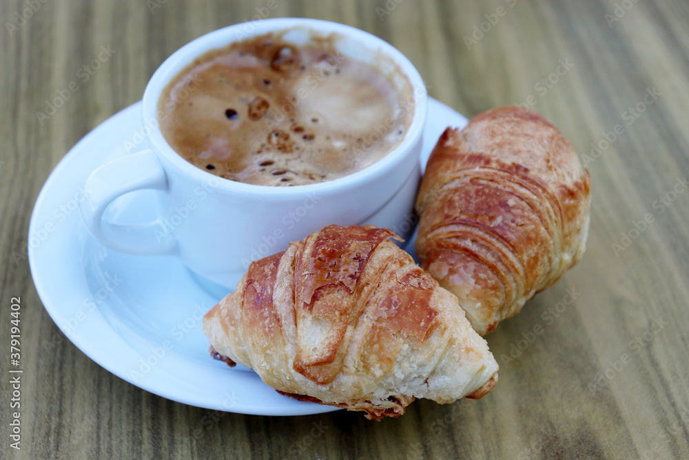 Cup of coffee and two croissants in a white saucer on wooden table. Tasty breakfast, cappuccino and fresh pastry close up Cup of coffee and two croissants in a white saucer on wooden table. Tasty breakfast, cappuccino and fresh pastry close up