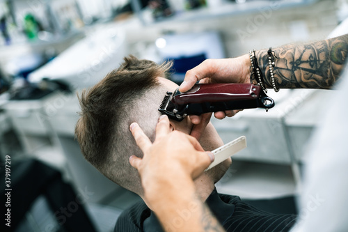 Selective focus of tattooed barber cutting client with hair clipper and holding comb in salon 