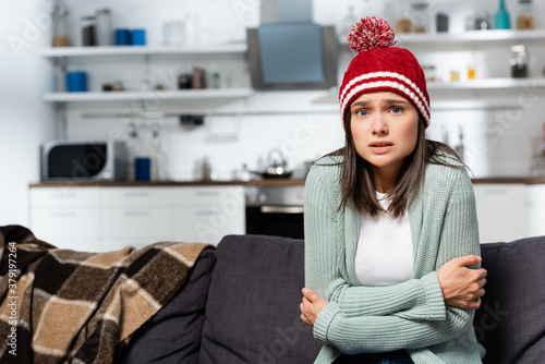 Obraz na plátně freezing woman in knitted hat hugging herself while sitting in cold kitchen