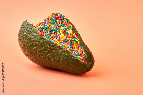 Avocado filling with sweet candy sprinklers. Close up green avocado full of multicolored sprinklers isolated on orange background. photo