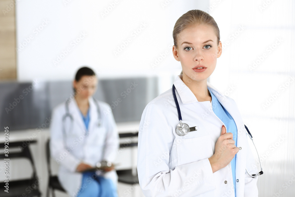 Woman - doctor standing in clinic with colleague at the background. Physician at work. Medicine and health care concept Woman - doctor standing in clinic with colleague at the background. Physician at work. Medicine and health care concept