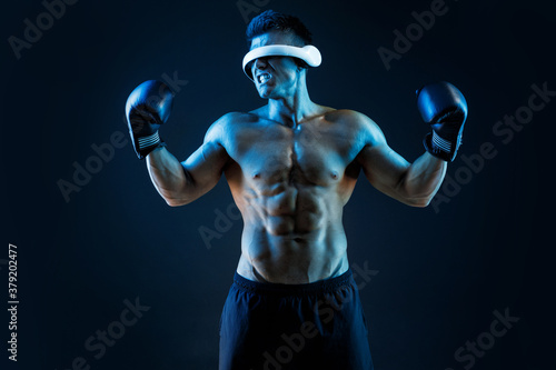 Muscular model sports young man in glasses of virtual reality and boxing gloves on dark background. Fashion portrait of strong brutal guy. Sexy torso. Male flexing his muscles. VR. Blue neon light. © KDdesignphoto