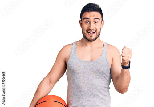 Young handsome man holding basketball ball screaming proud, celebrating victory and success very excited with raised arms © Krakenimages.com