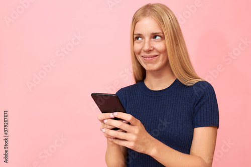Studio shot of beautiful 20 year old young woman with long blonde hair holding smart phone, flirting with cute guy via online dating app. Stylish teenage girl commenting posts on social media