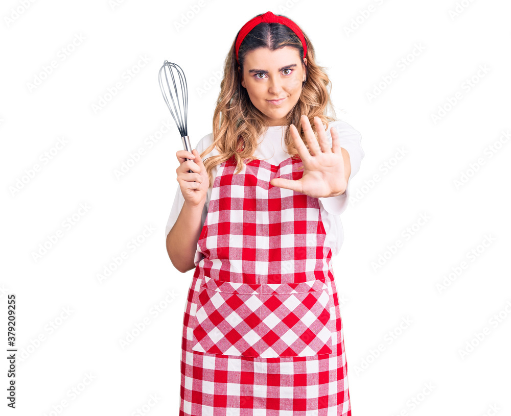 Young caucasian woman wearing apron holding whisk with open hand doing stop sign with serious and confident expression, defense gesture Young caucasian woman wearing apron holding whisk with open hand doing stop sign with serious and confident expression, defense gesture