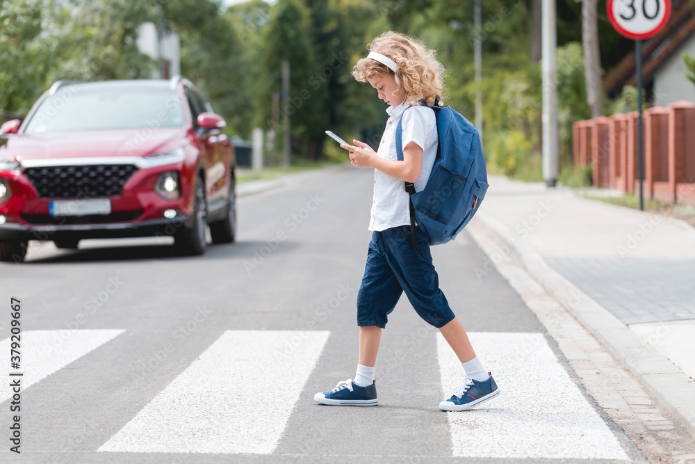 Adorable boy with a backpack, headphones and cellphone goes through the pedestrian crossing, not looking at red cars let him through