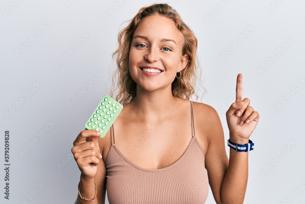 Beautiful caucasian woman holding birth control pills surprised with an idea or question pointing finger with happy face, number one Beautiful caucasian woman holding birth control pills surprised with an idea or question pointing finger with happy face, number one