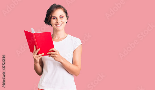Beautiful young woman with short hair reading a book looking positive and happy standing and smiling with a confident smile showing teeth