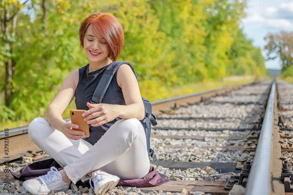 happy pretty red-haired young woman sitting on the railroad track with phone in her hands happy pretty red-haired young woman sitting on the railroad track with phone in her hands