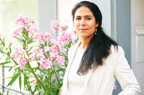 Beautiful middle age woman posing outside, wearing white jacket, standing next too pink flowers