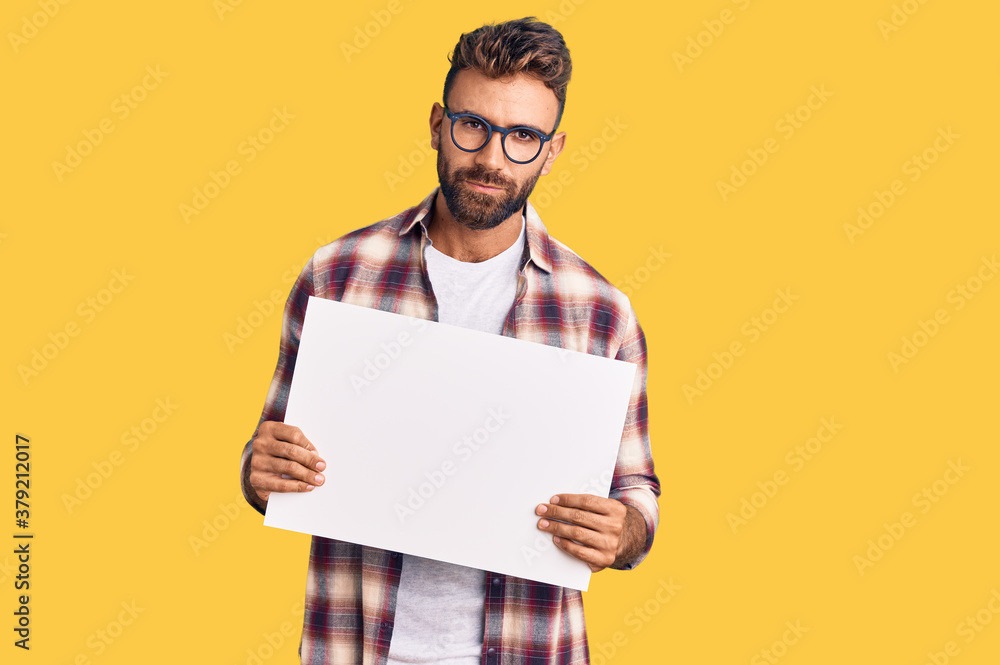 Young hispanic man holding blank empty banner thinking attitude and sober expression looking self confident Young hispanic man holding blank empty banner thinking attitude and sober expression looking self confident