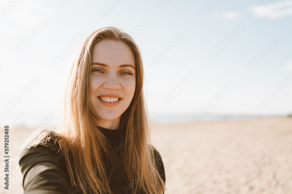 Pretty female with long blonde hair taking selfie on mobile phone on sandy beach in summer or autumn. Beautiful woman looking at camera and smiling in sunny day in ocean or sea coastline