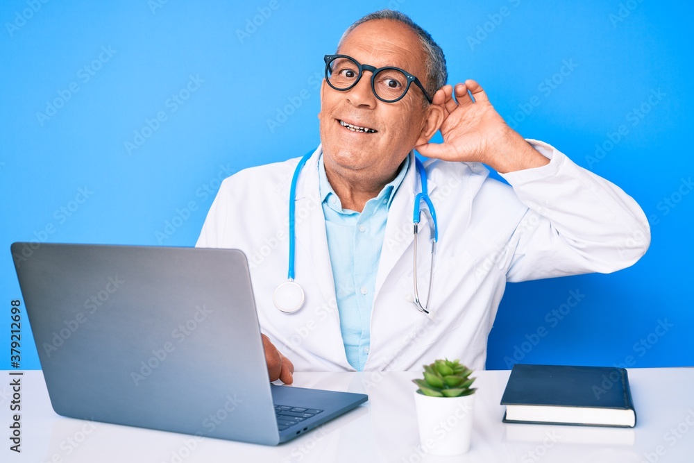 Senior handsome man with gray hair wearing doctor uniform working using computer laptop smiling with hand over ear listening an hearing to rumor or gossip. deafness concept. Senior handsome man with gray hair wearing doctor uniform working using computer laptop smiling with hand over ear listening an hearing to rumor or gossip. deafness concept.