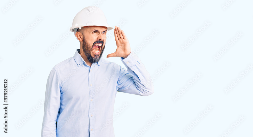 Young handsome man wearing architect hardhat shouting and screaming loud to side with hand on mouth. communication concept. Young handsome man wearing architect hardhat shouting and screaming loud to side with hand on mouth. communication concept.