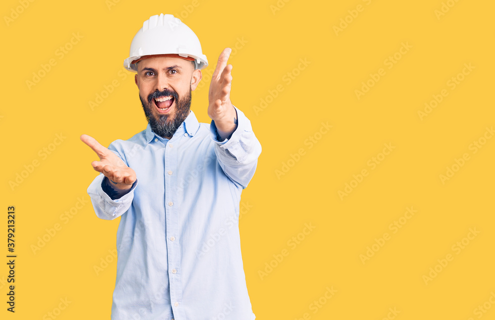 Young handsome man wearing architect hardhat looking at the camera smiling with open arms for hug. cheerful expression embracing happiness.