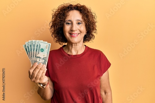 Beautiful middle age mature woman holding 20 dollars banknotes looking positive and happy standing and smiling with a confident smile showing teeth