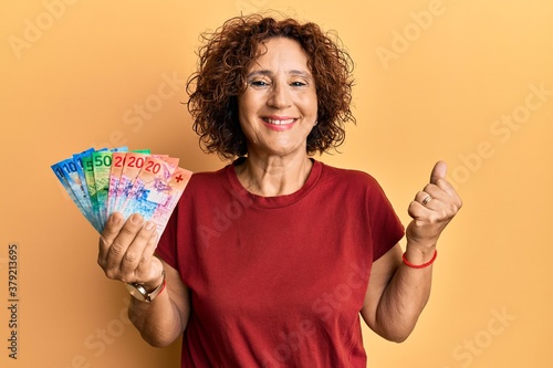 Beautiful middle age mature woman holding swiss franc banknotes screaming proud, celebrating victory and success very excited with raised arm